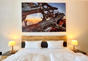 A hotel room with a large painting of a tree and sunset, a wooden headboard, two white pillows, and two bedside lamps. - Ostseeurlaub mit Stil - Moderne Ferienwohnung in Sellin (Sellin)