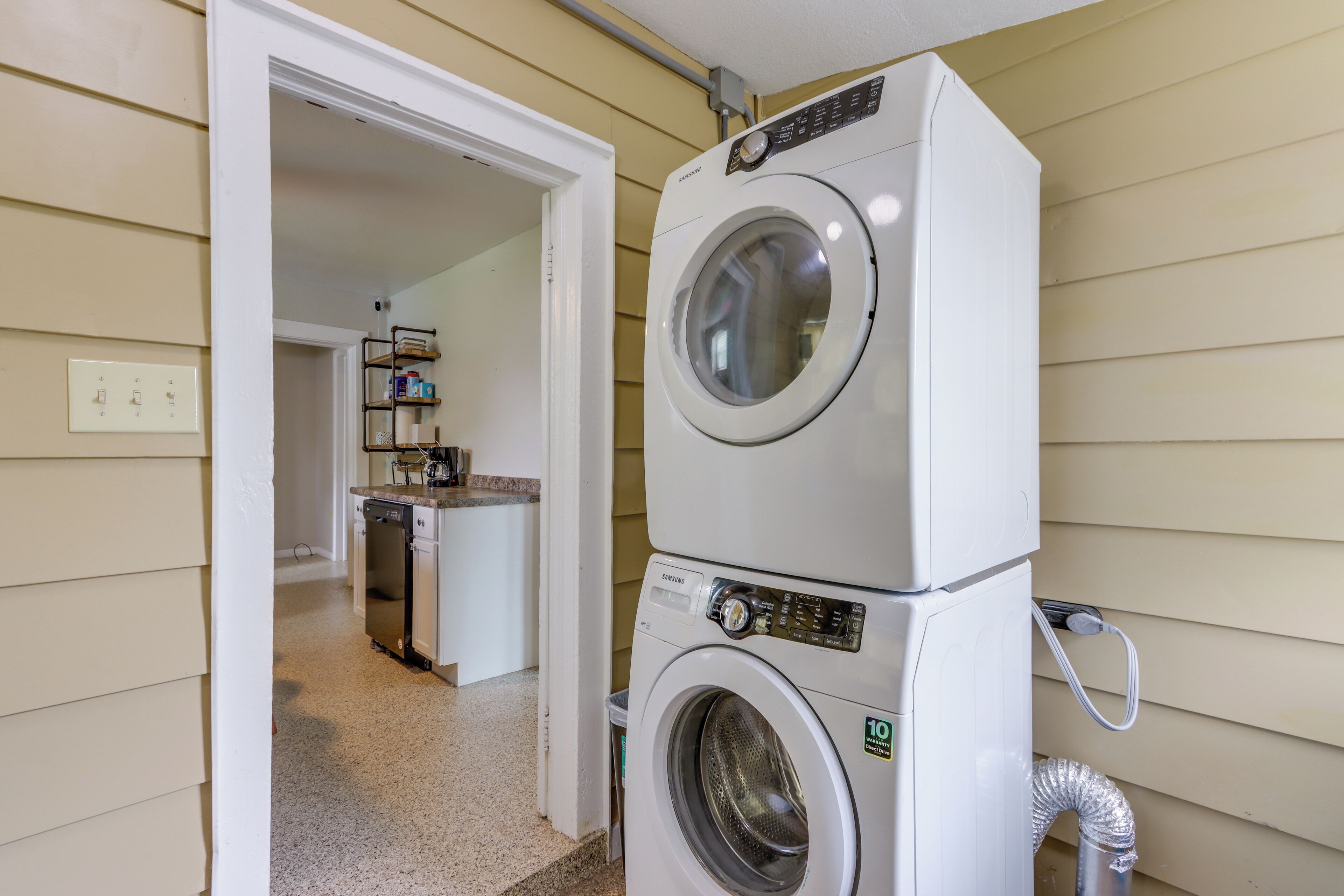 Une buanderie avec des lave-linge et des sèche-linge superposés, un coin cuisine à l’arrière-plan et une porte donnant sur une autre pièce.