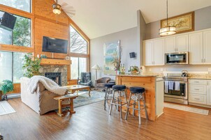 A spacious living area with a fireplace, wooden flooring, a large window, and a kitchen with white cabinets and a microwave.