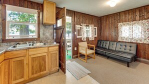 A kitchen with wooden cabinets, a built-in sink, and a window. There is a sofa and a chair in the living area. - Pet-friendly Bass Lake Cabin: Beach & Boat Dock (Irons)