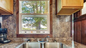 A kitchen with a double stainless steel sink, wooden cabinets, a window with a view of trees, and a coffee maker on the counter. - Pet-friendly Bass Lake Cabin: Beach & Boat Dock (Irons)