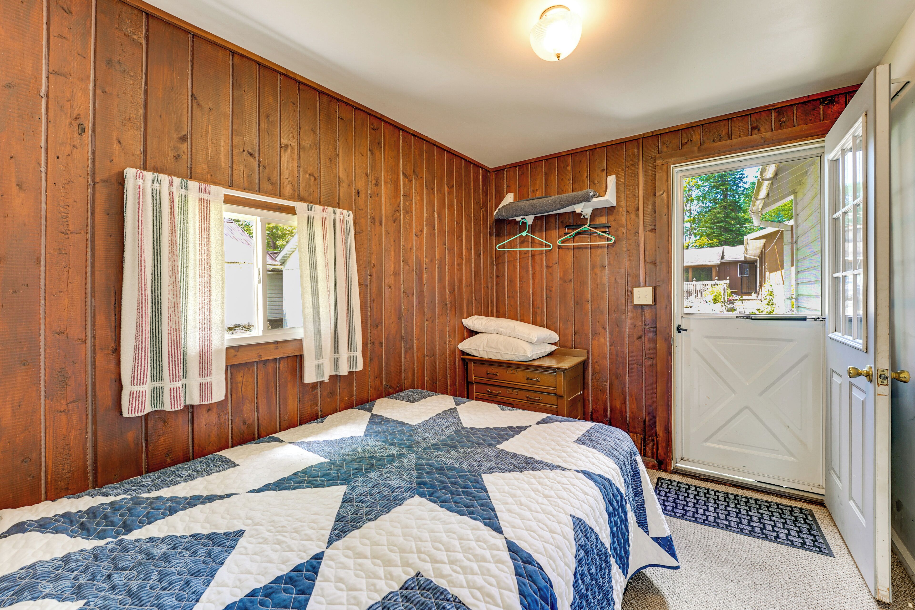 Une chambre avec un mur en bois, un lit recouvert d’une courtepointe, une fenêtre avec des rideaux, une porte et une petite table avec un coussin.