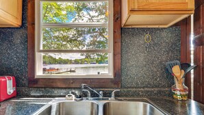A kitchen with a double stainless steel sink, a window with a view of a lake, and wooden cabinets. - Swim, Boat, Fish On-site: Quaint Bass Lake Cabin! (Irons)