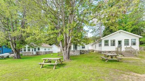 A white cabin with a stone foundation, surrounded by a grassy yard with picnic tables. - Swim, Boat, Fish On-site: Quaint Bass Lake Cabin! (Irons)