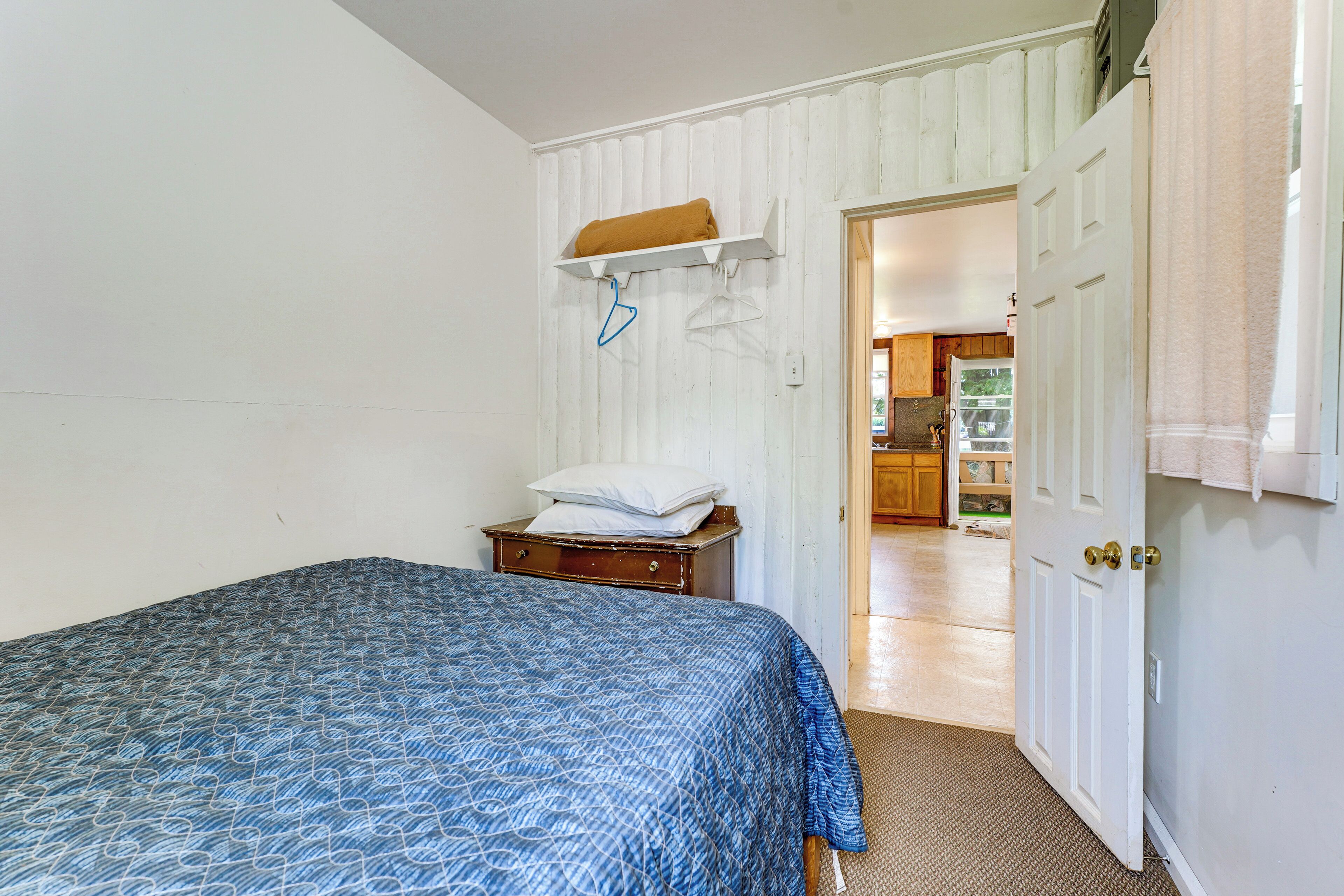 A bedroom with a bed, a wooden nightstand, a hanging towel rack, and a door leading to a kitchen.