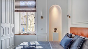 A bedroom with a bed, a window with blinds, a wall-mounted light, and a decorative sculpture. - New! Luxury Old town apartment (Vilnius)