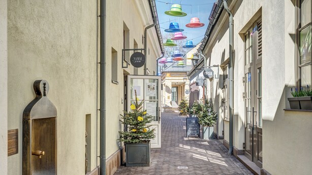 A narrow alleyway with a herringbone brick floor, a visible restroom on the left, and a shop with a signboard on the right. - New! Luxury Old town apartment (Vilnius)