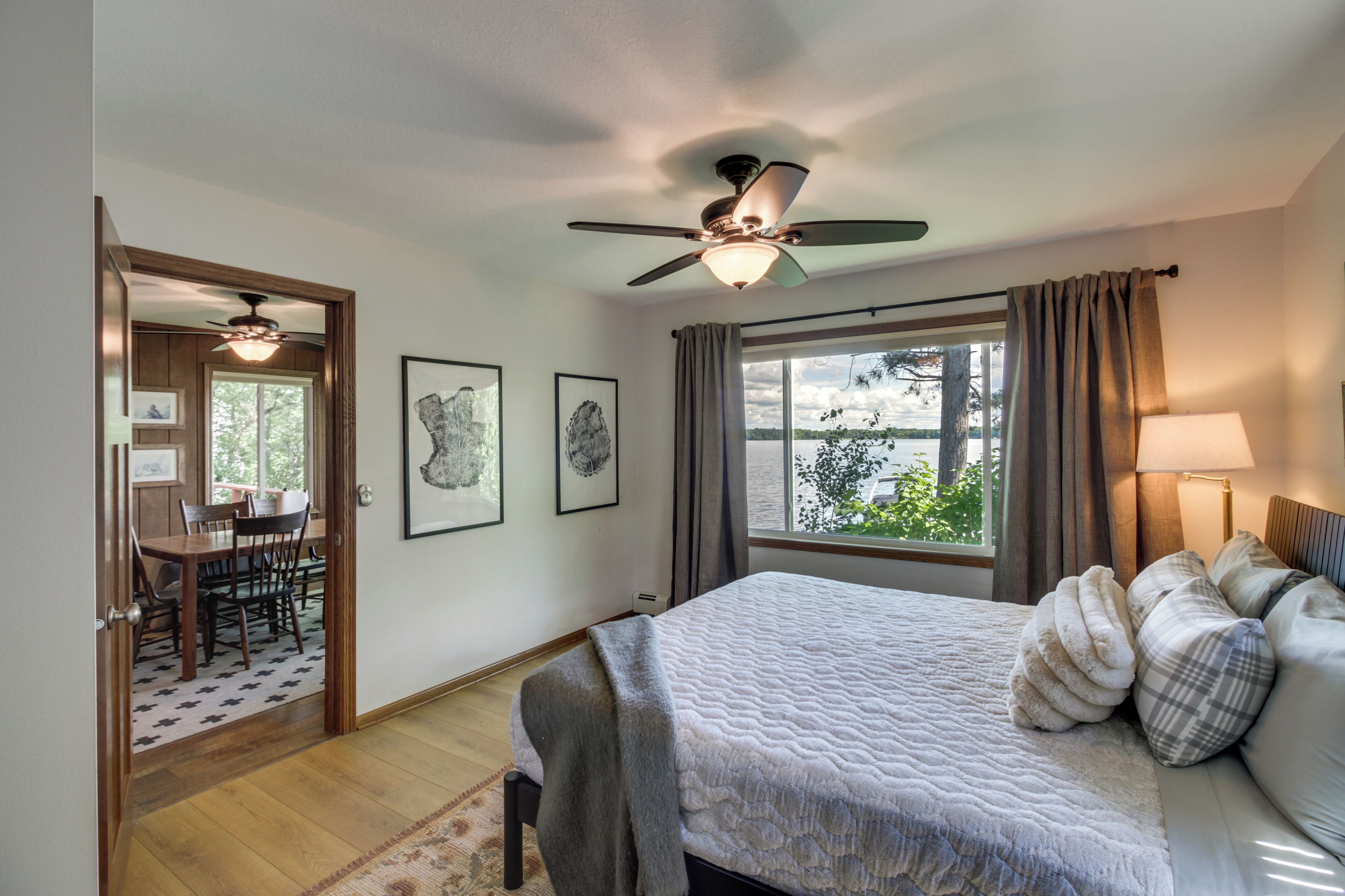 A bedroom with a bed, a ceiling fan, a window with curtains, a dining area with a table and chairs, and framed artwork on the wall.