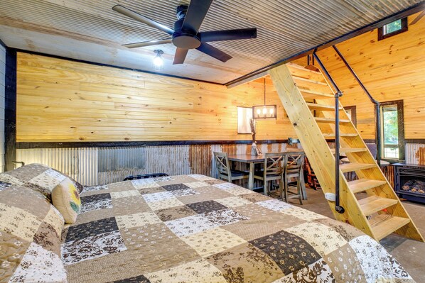 A bedroom with a wooden ceiling, a quilted bedspread, a ceiling fan, a wooden ladder, and a fireplace.