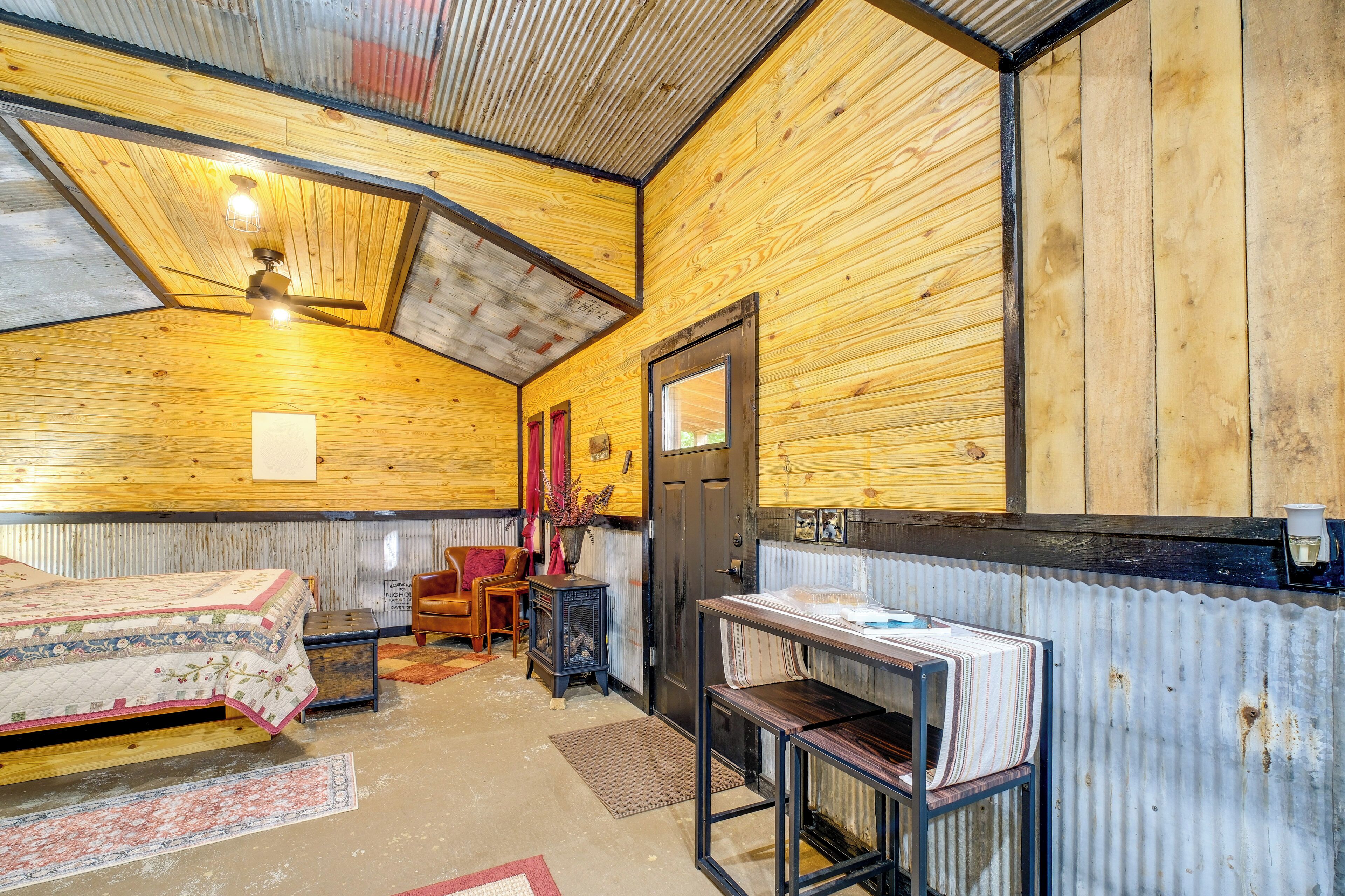 A rustic cabin interior with a wooden ceiling, a bed, a table, a chair, and a fireplace.