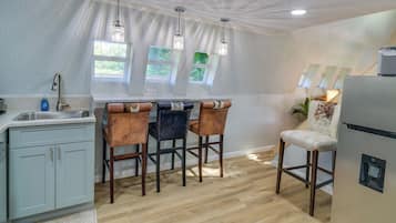 A kitchen with a bar area, wooden chairs, a refrigerator, and a window with a view of greenery.