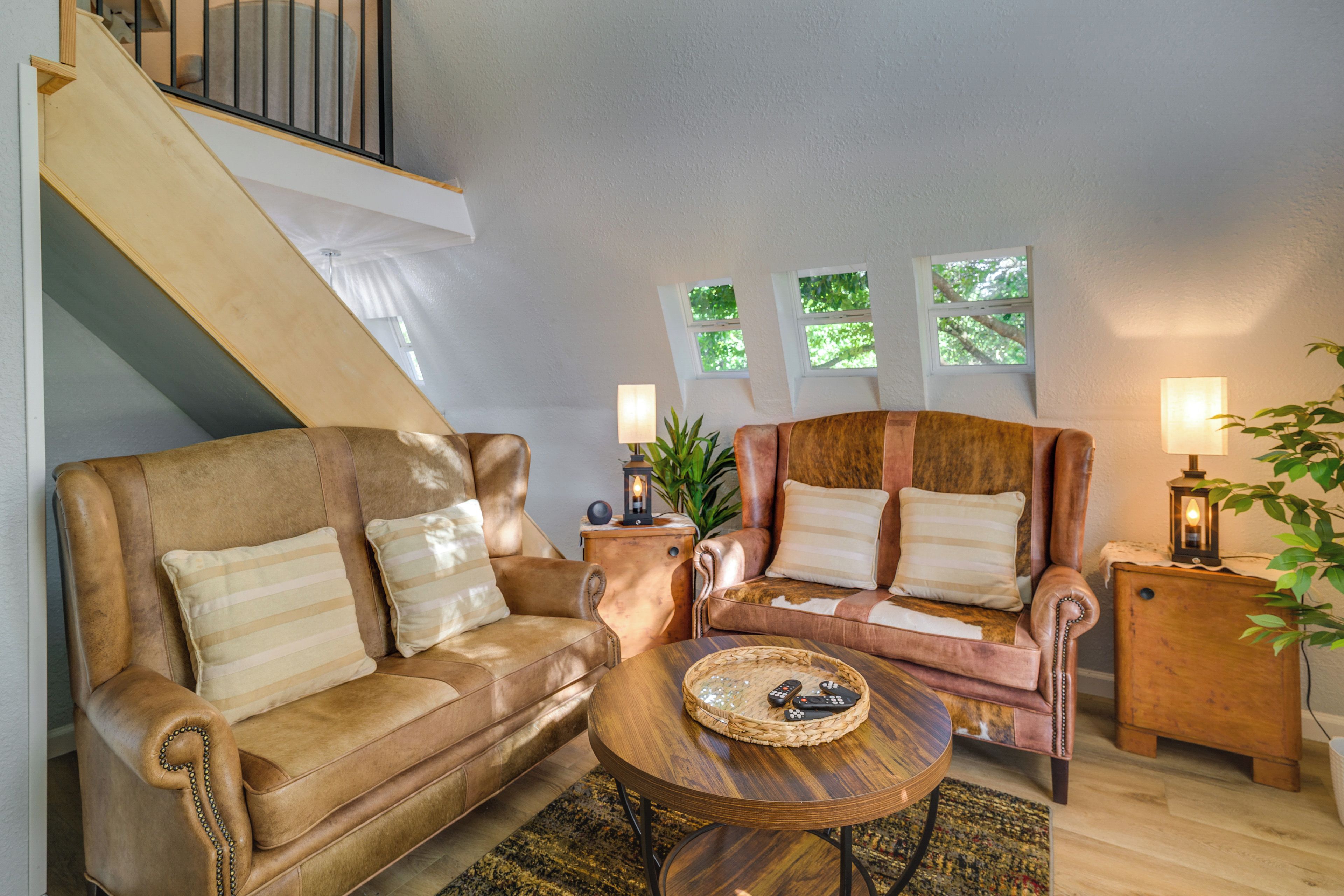 A living room with a sofa, armchair, and a coffee table. There is a staircase and a window with a view of trees.