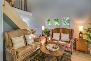 Una sala de estar con un sofá, un sillón y una mesa de centro. Se ve una escalera y una ventana con vista a árboles.