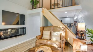 Una sala de estar con un sofá, una mesa de centro con controles remotos, un televisor montado en la pared y una escalera que sube a un nivel superior.