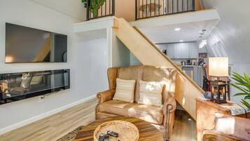 A living room with a sofa, a coffee table with remotes, a TV mounted on the wall, and a staircase leading to an upper level.