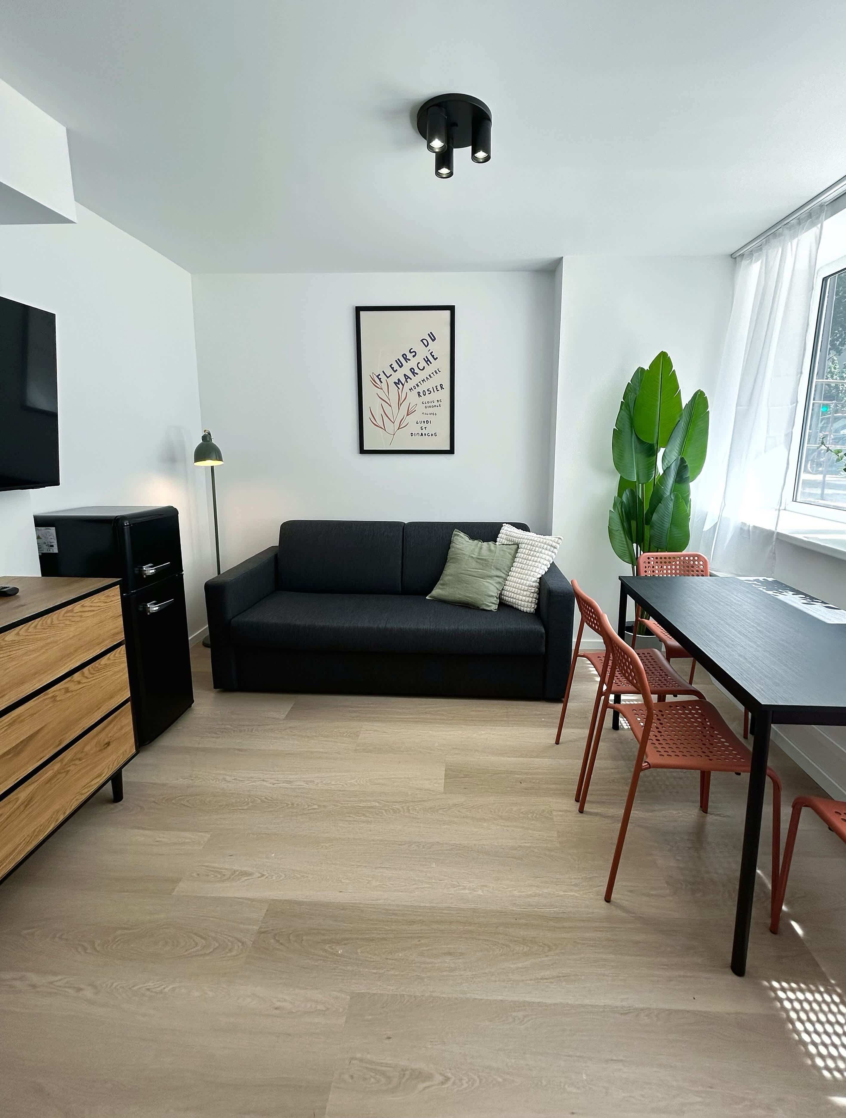 En moderne stue med en sort sofa, en trækommode, et spisebord med stole, en stor stueplante og et indrammet kunstværk på væggen.