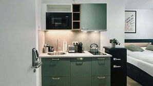 Een kleine keuken met een magnetron, spoelbak en kastjes, direct naast een bed met hoofdbord en nachtkastje.