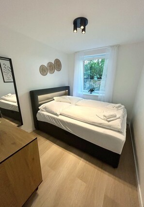 A neatly made bed with white linens, a bedside table, a window with a view of greenery, and a wall-mounted light fixture. - Modern Apartment in Eimsbuettel Hamburg for 5 (Hamburg)