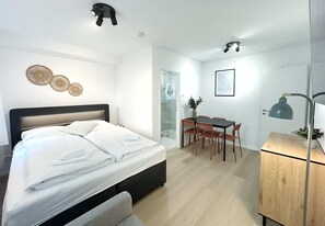 A bedroom with a bed, a desk, and a chair, a small cabinet, and a wall-mounted lamp.