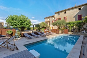 Une piscine avec des chaises longues, un mur en pierre et un bâtiment aux volets rouges.