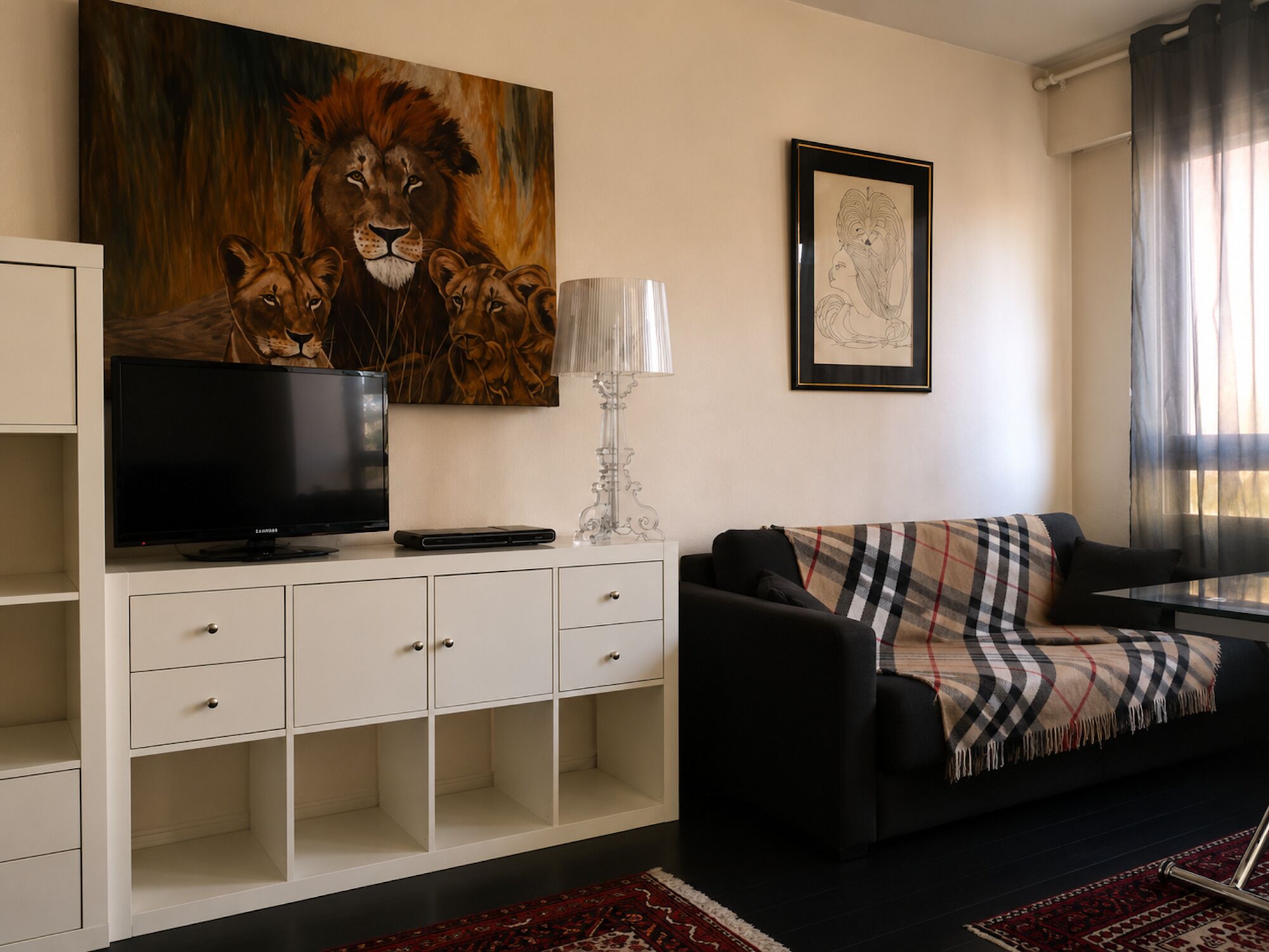 A living room with a television, a painting of lions, a lamp, and a couch with a plaid blanket.