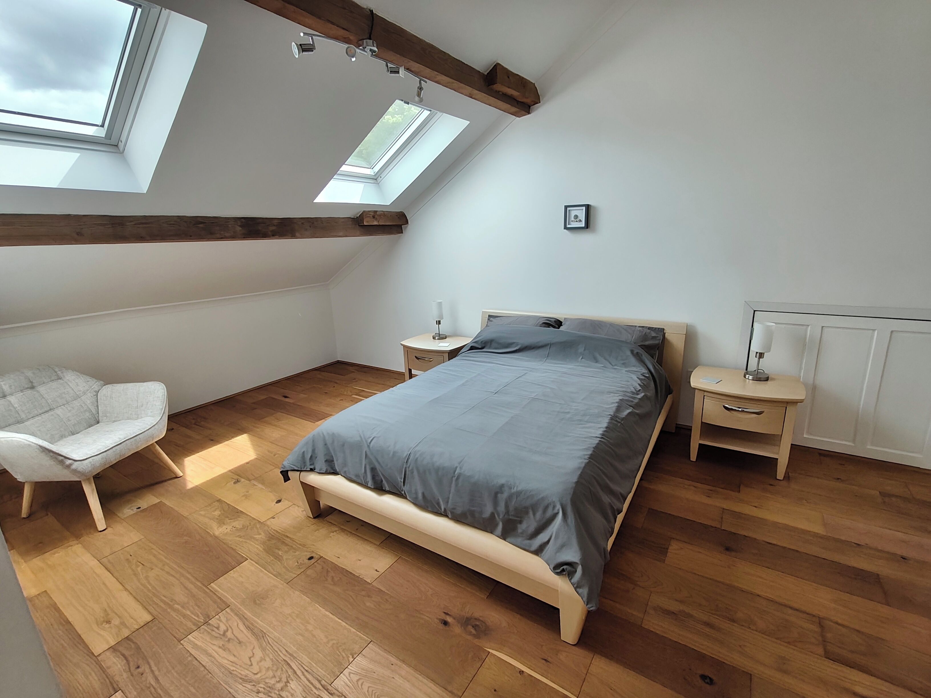 Un dormitorio con techo inclinado, tragaluces, vigas de madera, una cama, una silla, mesitas de noche y un radiador.