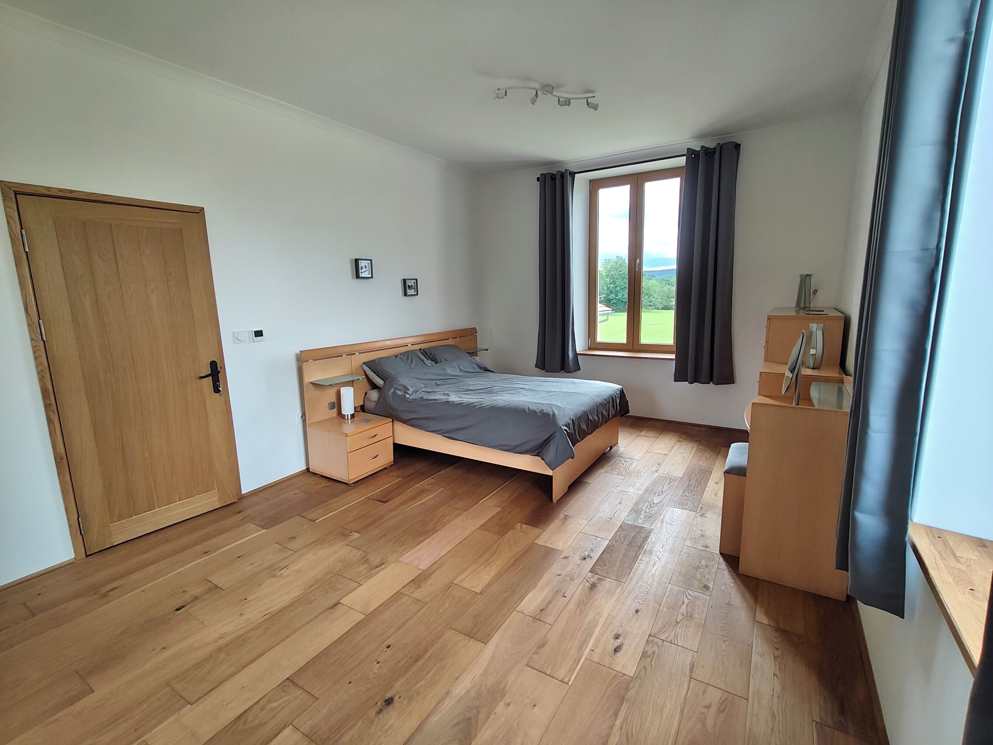 Une chambre avec un plancher en bois, un lit, une table de chevet, une commode, une fenêtre avec des rideaux et une porte.