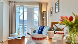 A hotel room with a dining table, a sofa, a coffee table, a window with curtains, and a view of the outside. - Moderne Ferienwohnung mit Balkon in Sudostlage in Sellin (Sellin)