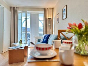 Ein Hotelzimmer mit Esstisch, einer Couch, einem Couchtisch, einem Fenster mit Vorhängen und Blick nach draußen.
