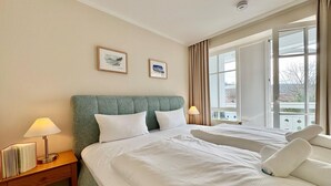A hotel room with two beds, a nightstand with a lamp, a window with curtains, and two framed pictures on the wall. - Moderne Ferienwohnung mit Balkon in Sudostlage in Sellin (Sellin)