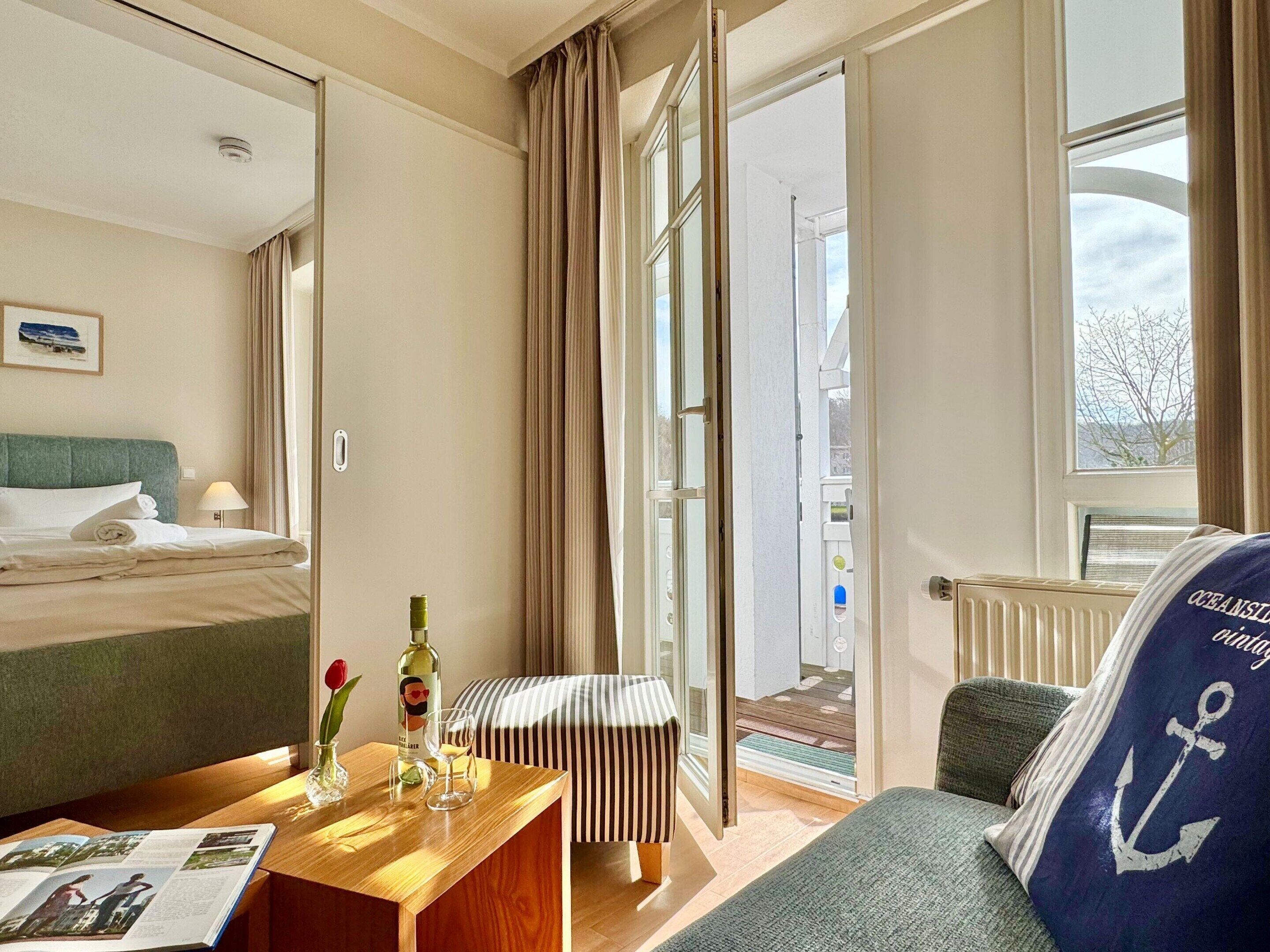 Une chambre d’hôtel avec un lit, un bureau sur lequel se trouve un magazine, une bouteille d’alcool, un verre et un vase avec une fleur.
