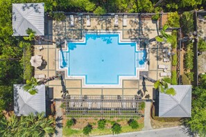 Ein Luftbild zeigt einen rechteckigen Swimmingpool mit einer umlaufenden Liegefläche, beschattet durch große Sonnenschirme und umgeben von Palmen.