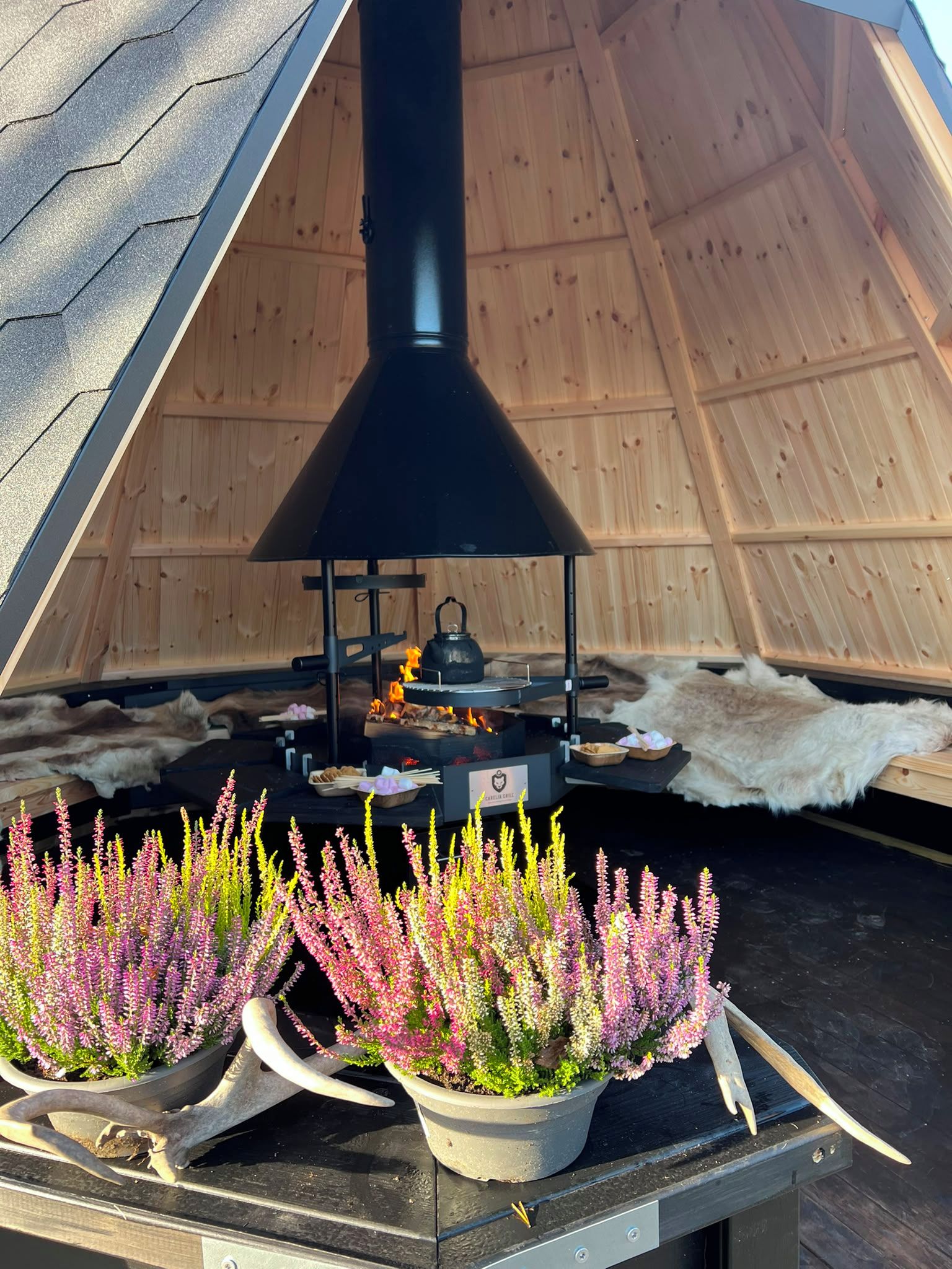 An outdoor cooking area with a black chimney, a kettle, and a fire burning. There are potted plants with pink flowers in front of the cooking area.
