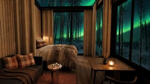 Una habitación acogedora con cama, sofá, una mesa con velas y vistas a un bosque a través de amplias ventanas.