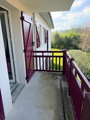 Ein überdachter Balkon mit roter Geländer, einer weißen Tür und Blick auf eine grüne Landschaft.