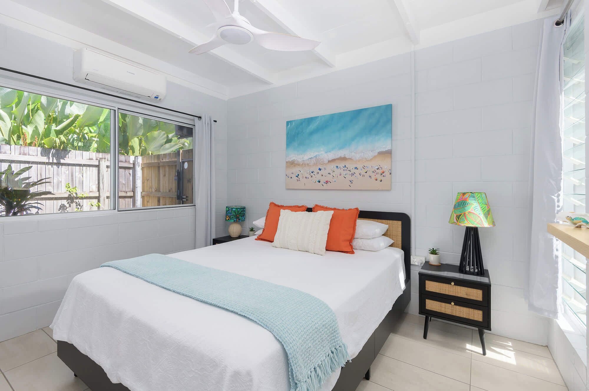 A bedroom with a bed, a nightstand, a lamp, a painting, and a window with a view of plants.