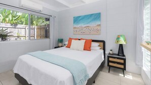 A bedroom with a bed, a nightstand, a lamp, a painting, and a window with a view of plants. - Paperbark Cottage - New Tropical Holiday Rental (Port Douglas)