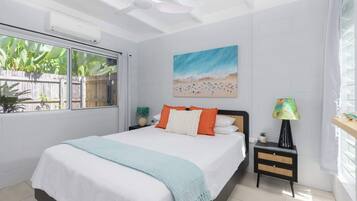 A bedroom with a bed, a nightstand, a lamp, a painting, and a window with a view of plants.