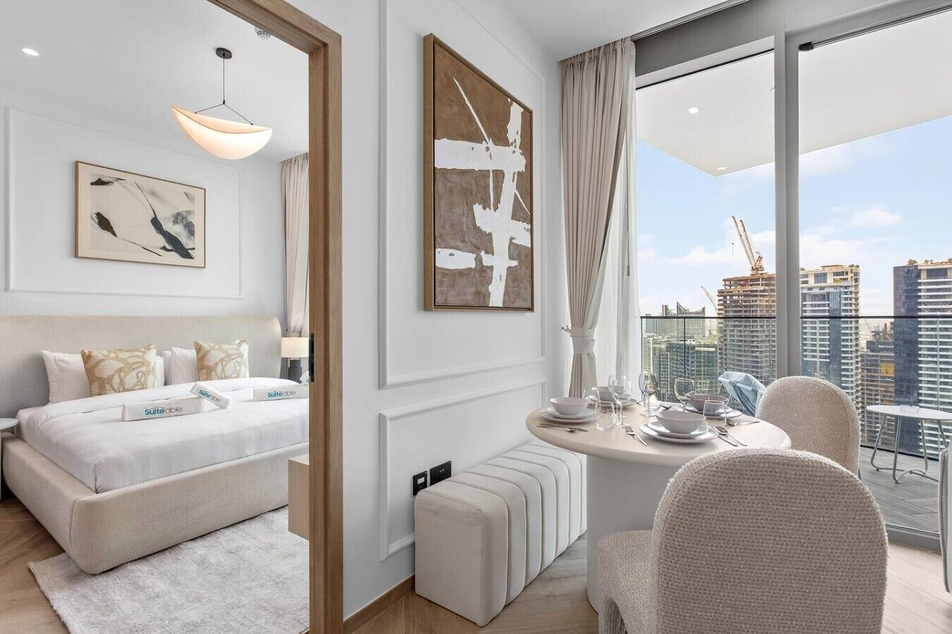 A modern hotel room with a bed, a dining table, and a view of the cityscape.