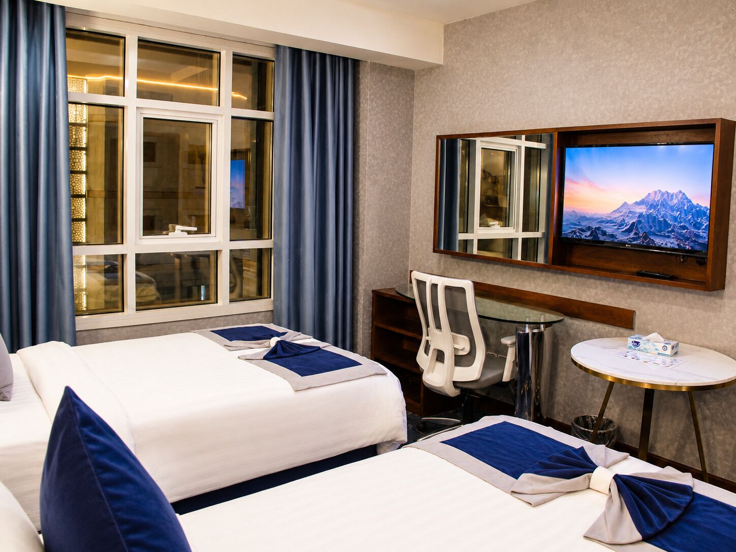A hotel room with two beds, a desk, a chair, a TV, and a small table with a tissue box.