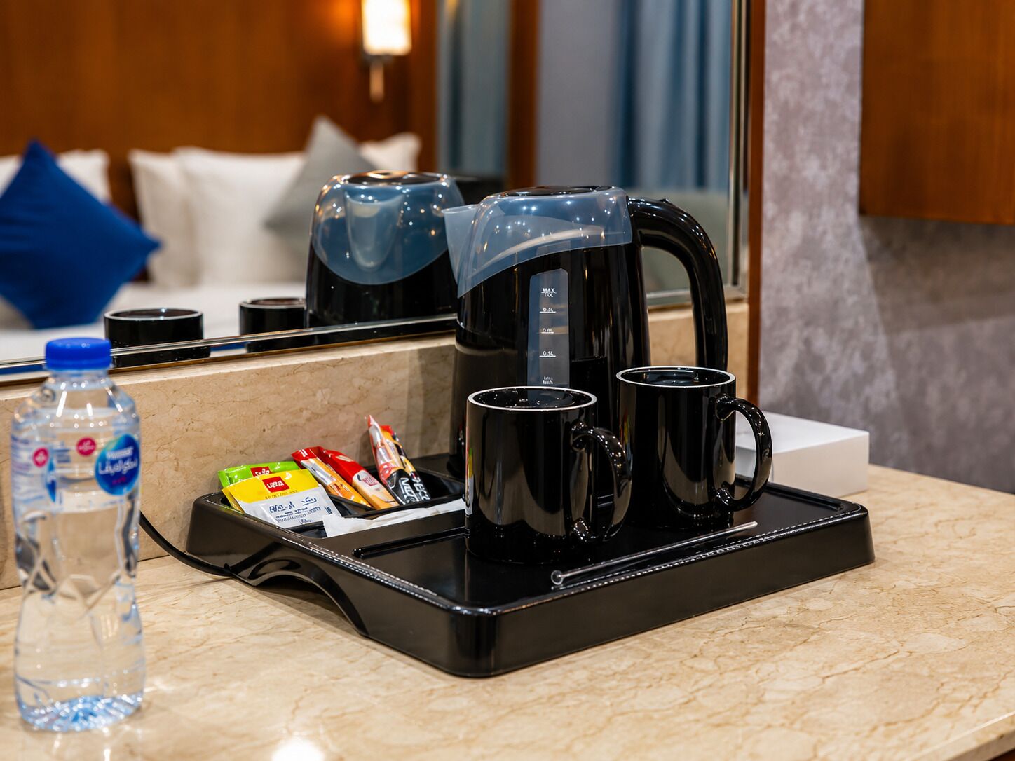 A coffee maker with two mugs on a tray, a water bottle, and some food packets on a countertop.
