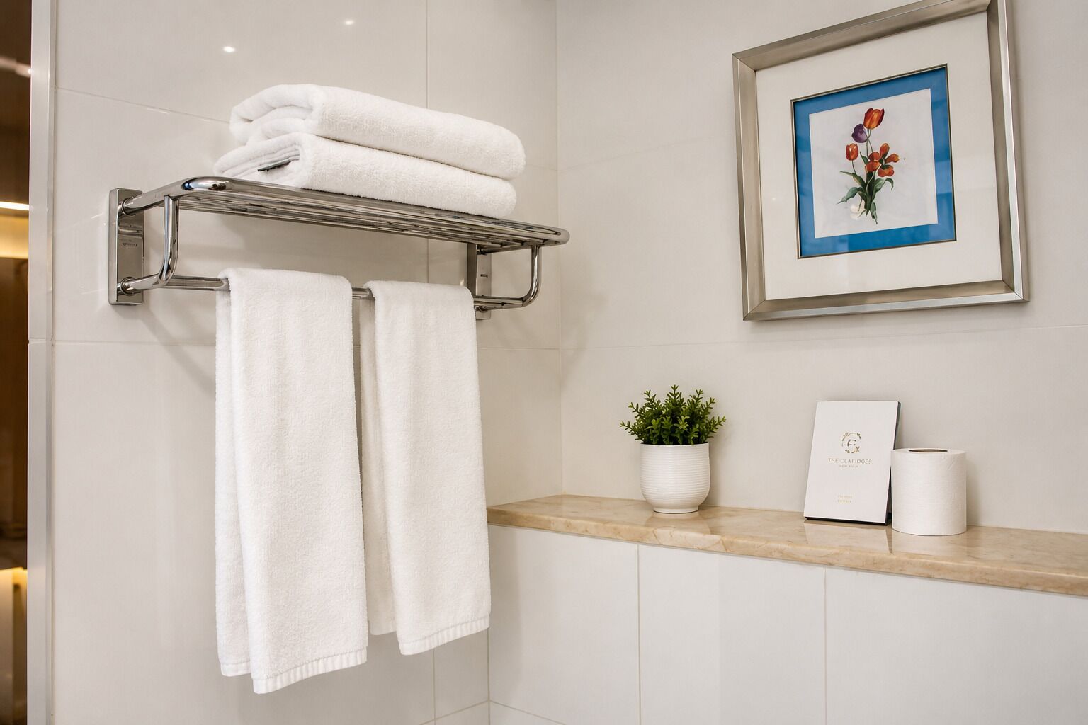 Ein Badezimmer mit Handtuchhalter, der zwei Handtücher trägt, einem gerahmten Bild von Blumen, einer Topfpflanze und einer Toilettenpapierrolle auf einem Regal.