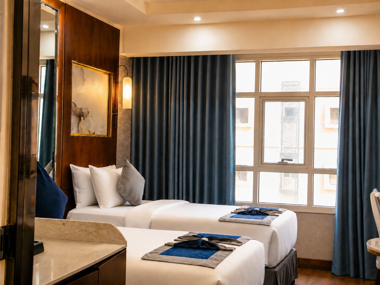 A hotel room with two beds, a marble bedside table, a painting on the wall, and a view through the window.