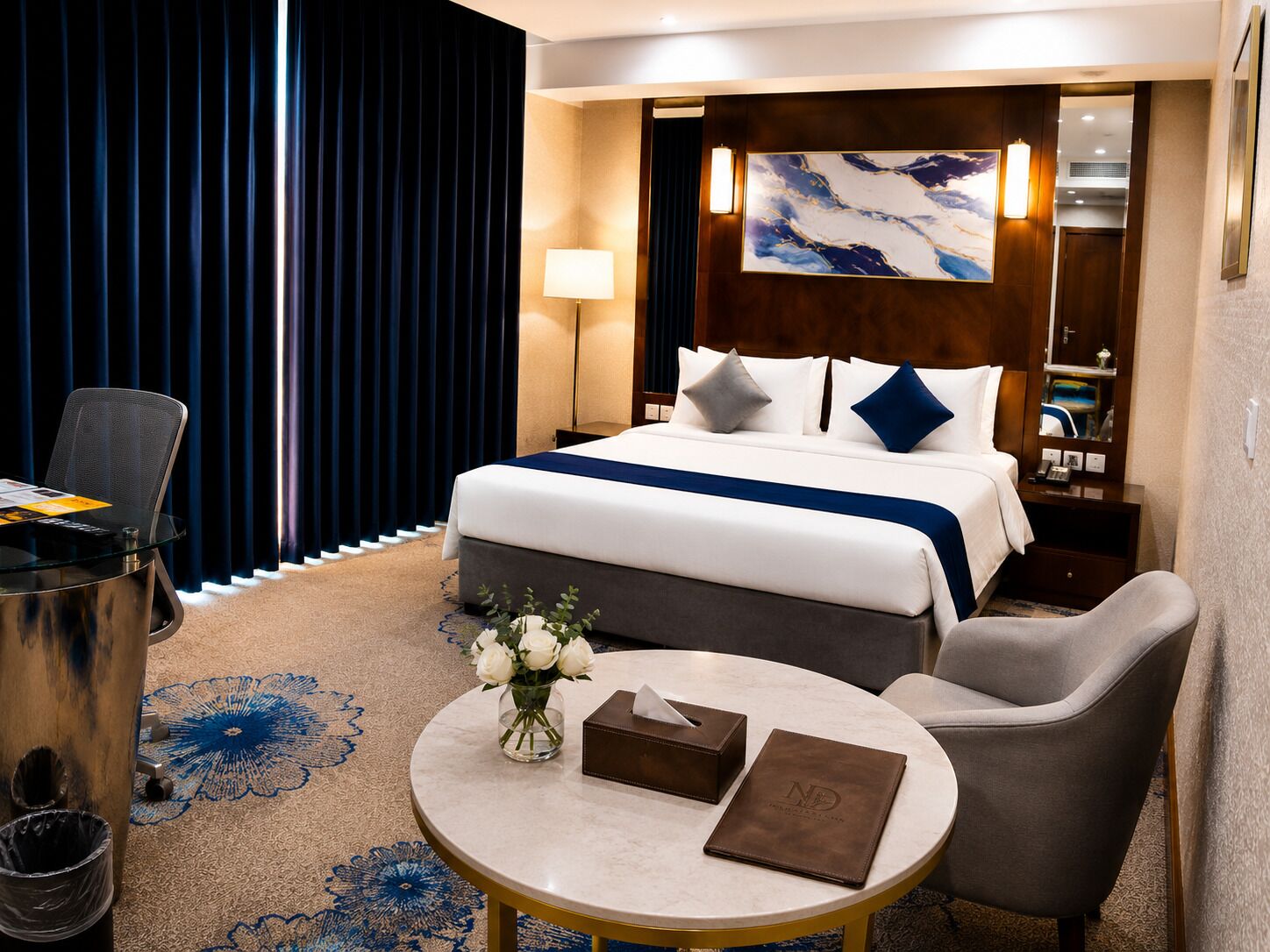 A hotel room with a bed, a desk, a chair, a small table with a vase of flowers, and a nightstand with a lamp.