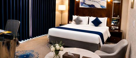 A hotel room with a bed, a desk, a chair, a small table with a vase of flowers, and a nightstand with a lamp.