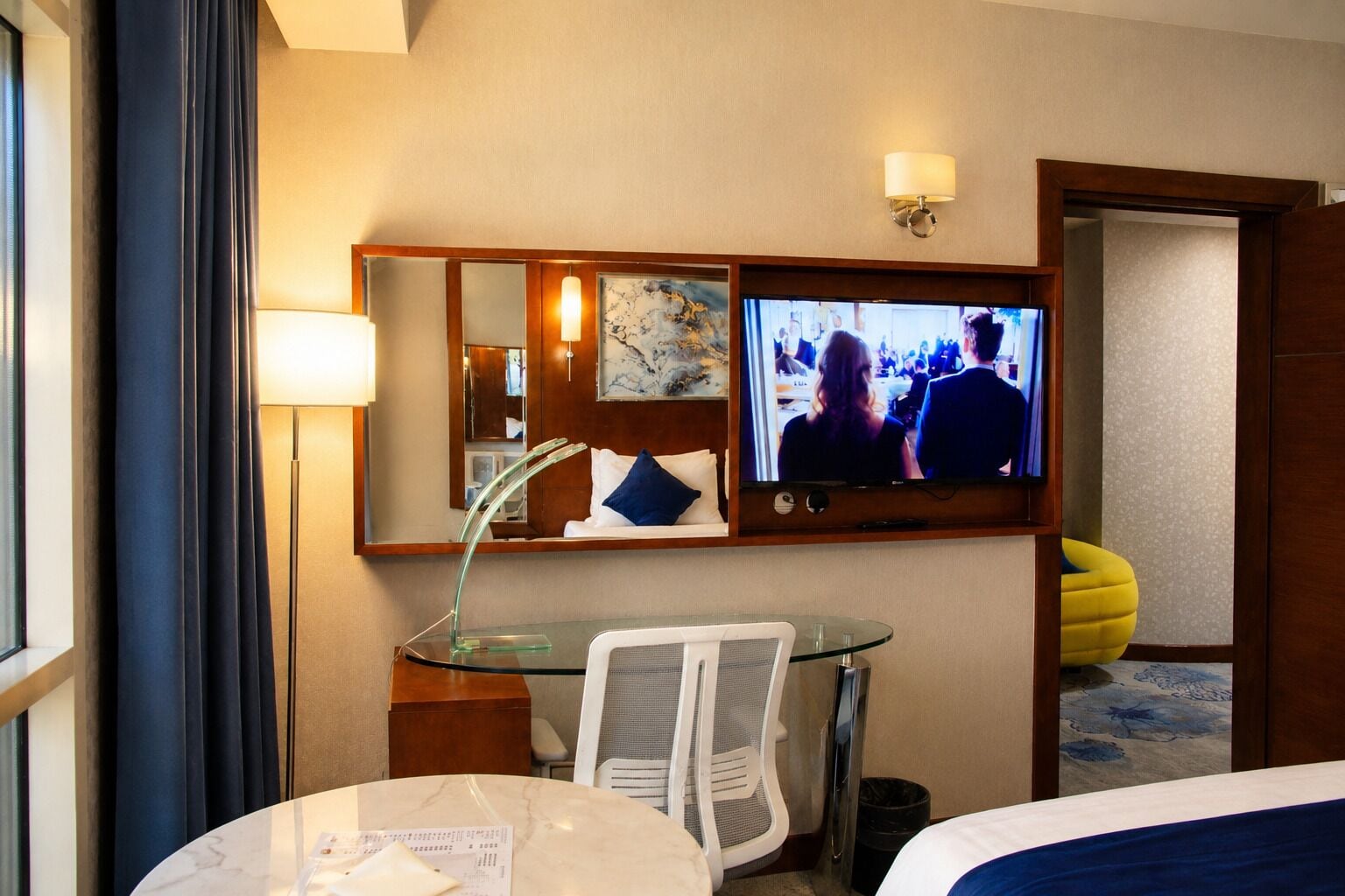 A hotel room with a bed, a desk with a chair, a television, a mirror, and a window with curtains.