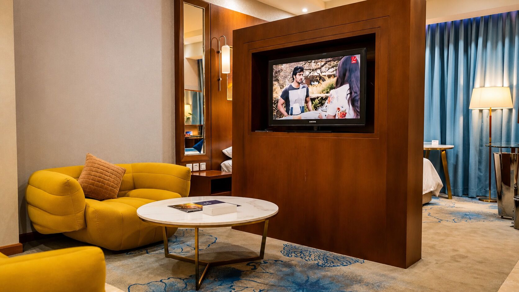 Quarto de hotel com poltrona amarela, mesa de centro redonda, televisão fixada em parede de madeira e cortina azul.