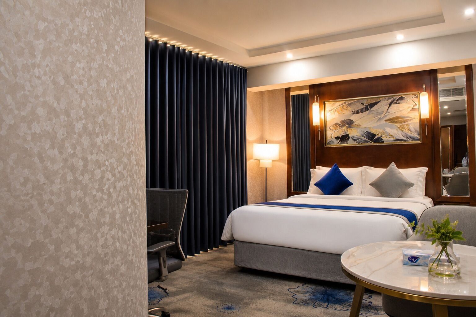 A hotel room with a bed, a desk, a chair, a round table with a vase, and a large painting on the wall.