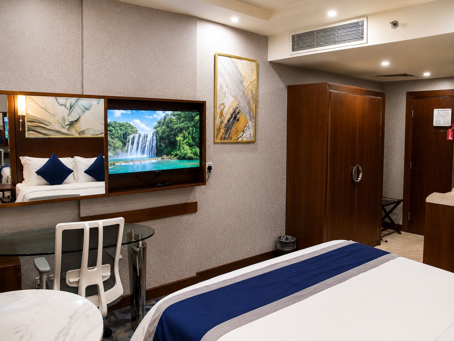 A hotel room with a bed, a TV mounted on the wall, a desk with a chair, and a wardrobe.