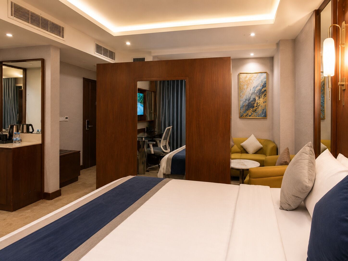A hotel room with a bed, a desk, a sofa, a TV, and a painting on the wall.